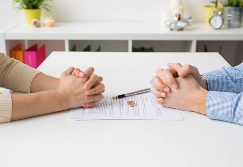 A couple sits with clenched hands across a table containing a divorce document and rings