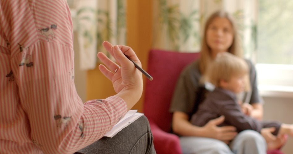 A therapist holds a pencil while a mother and child sit blurred behind
