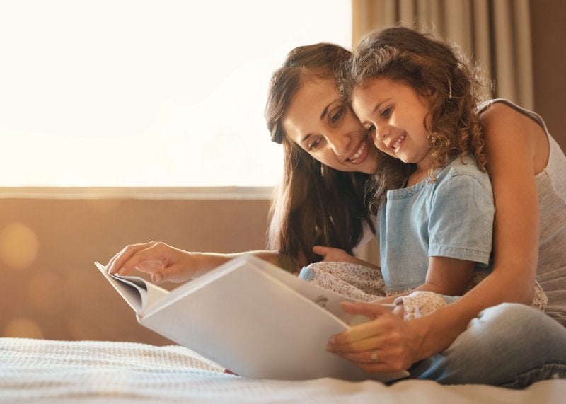 A mother and daughter smile while reading a book together on a bed