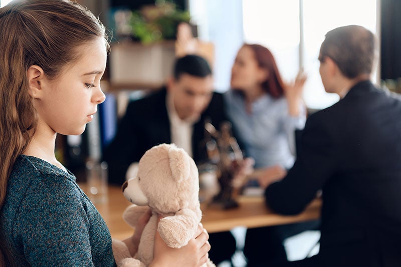 A sad young girl holds a teddy bear while adults argue behind her.